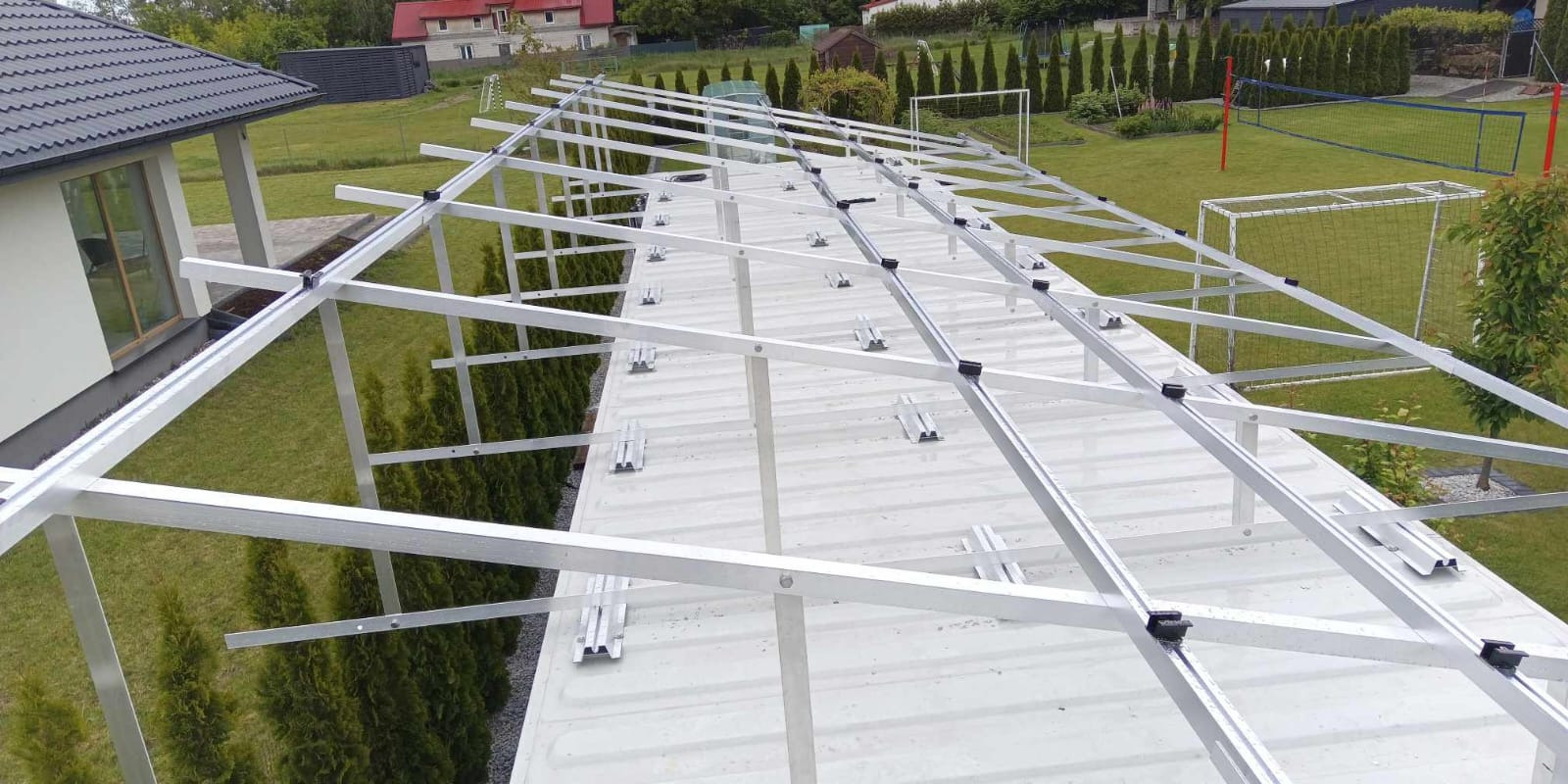 Montaż aluminiowej konstrukcji wsporczej na dachu pokrytym blachą trapezową, przygotowanej pod instalację paneli słonecznych. Widok z góry na tle zieleni.