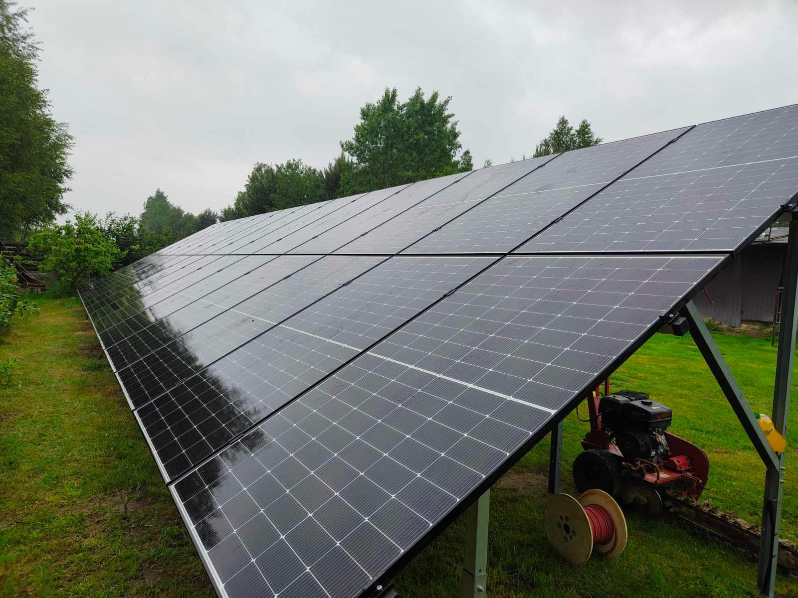 Ukończona instalacja fotowoltaiczna na trawniku, widoczna maszyna do kopania rowów i szpula z kablem. Pochmurne niebo w tle.