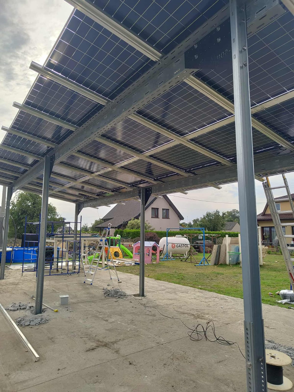 Montaż paneli słonecznych na metalowej konstrukcji w ogrodzie, widoczne elementy instalacji i otoczenie domu. Etap prac z widocznym okablowaniem i narzędziami.