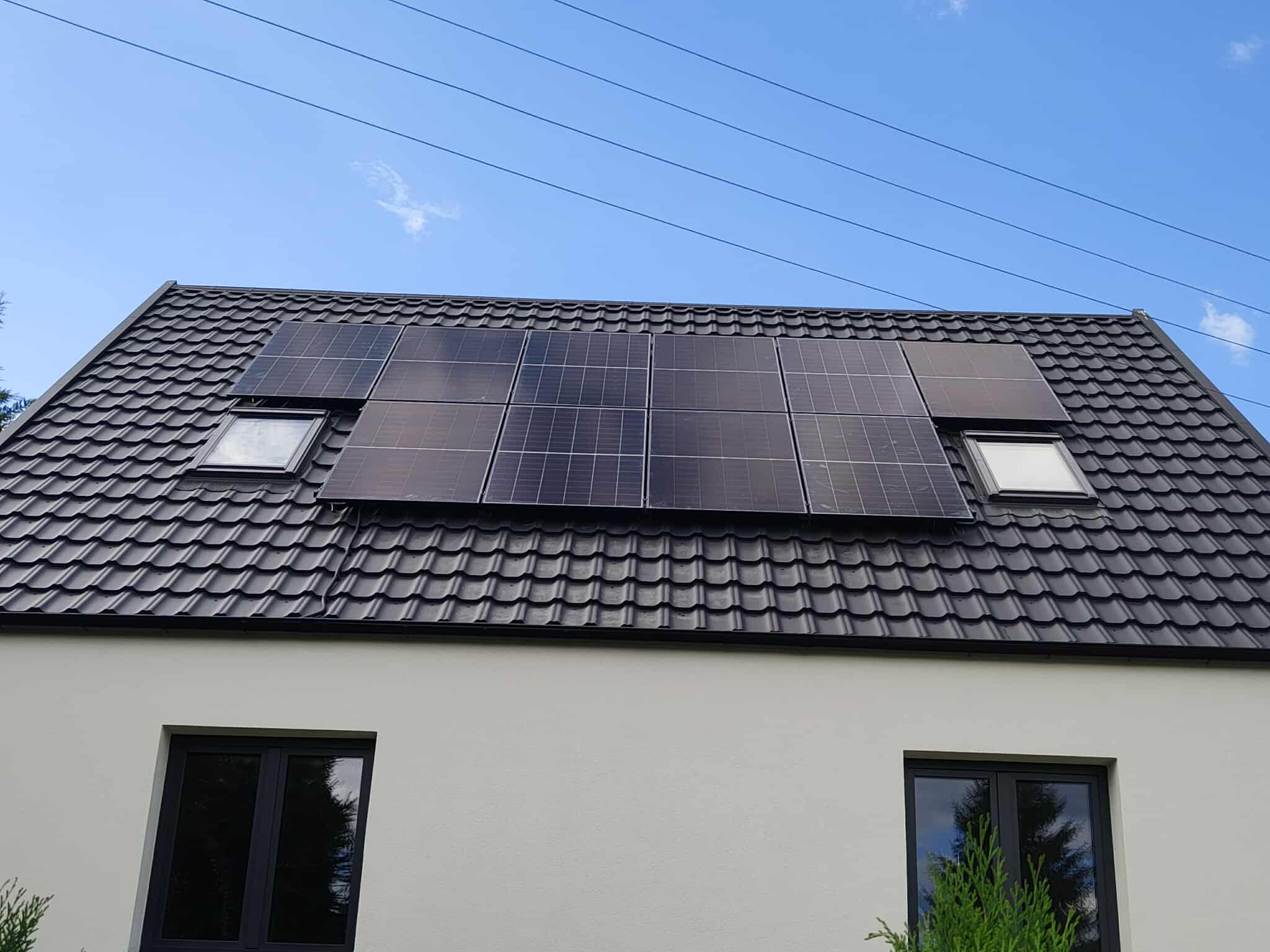 Panele fotowoltaiczne na dachu domu z dachówką w kolorze grafitowym, widoczne okna dachowe i fragment elewacji w jasnym kolorze. Błękitne niebo w tle.