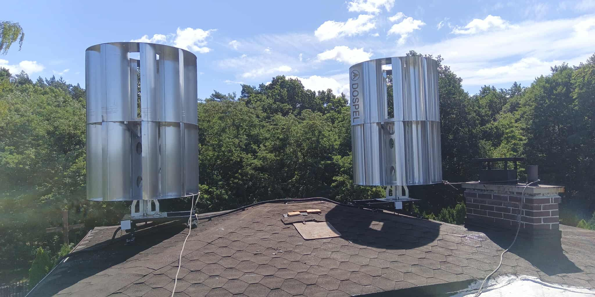 Dwie pionowe turbiny wiatrowe marki Dospel zamontowane na dachu domu z kominem, otoczone zielenią drzew i błękitnym niebem z chmurami.