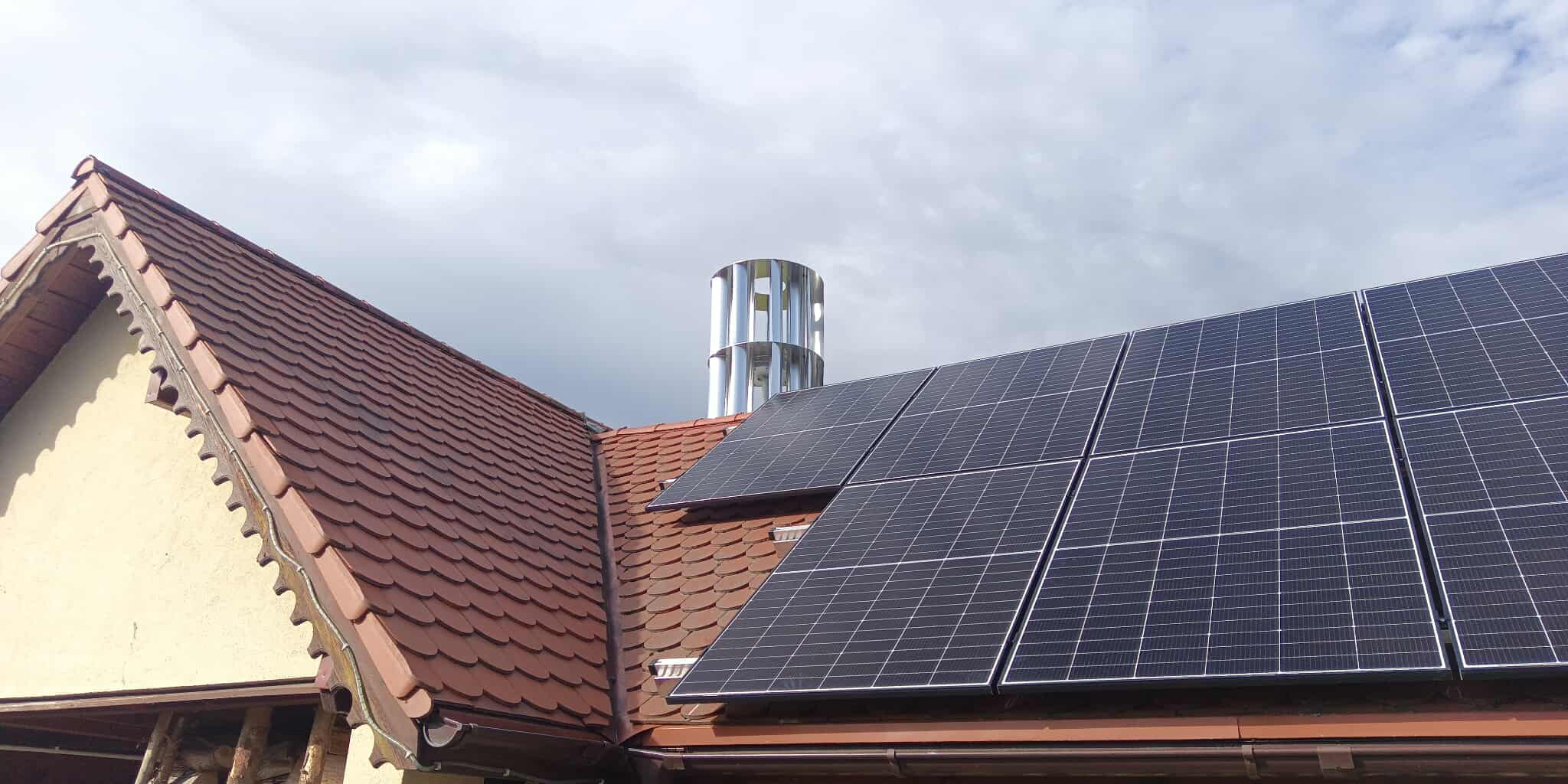 Czerwony dach z panelami słonecznymi i turbiną wiatrową w tle, widok z dołu. Panele zamontowane na połaci dachu, turbina wiatrowa w formie cylindrycznej konstrukcji.