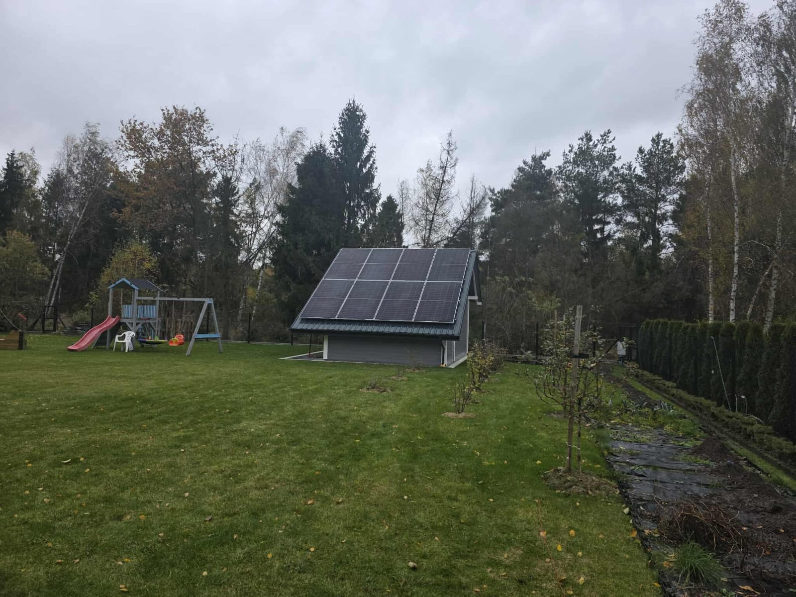Mały budynek z panelami słonecznymi na dachu, otoczony zielonym trawnikiem i drzewami. W tle plac zabaw dla dzieci.