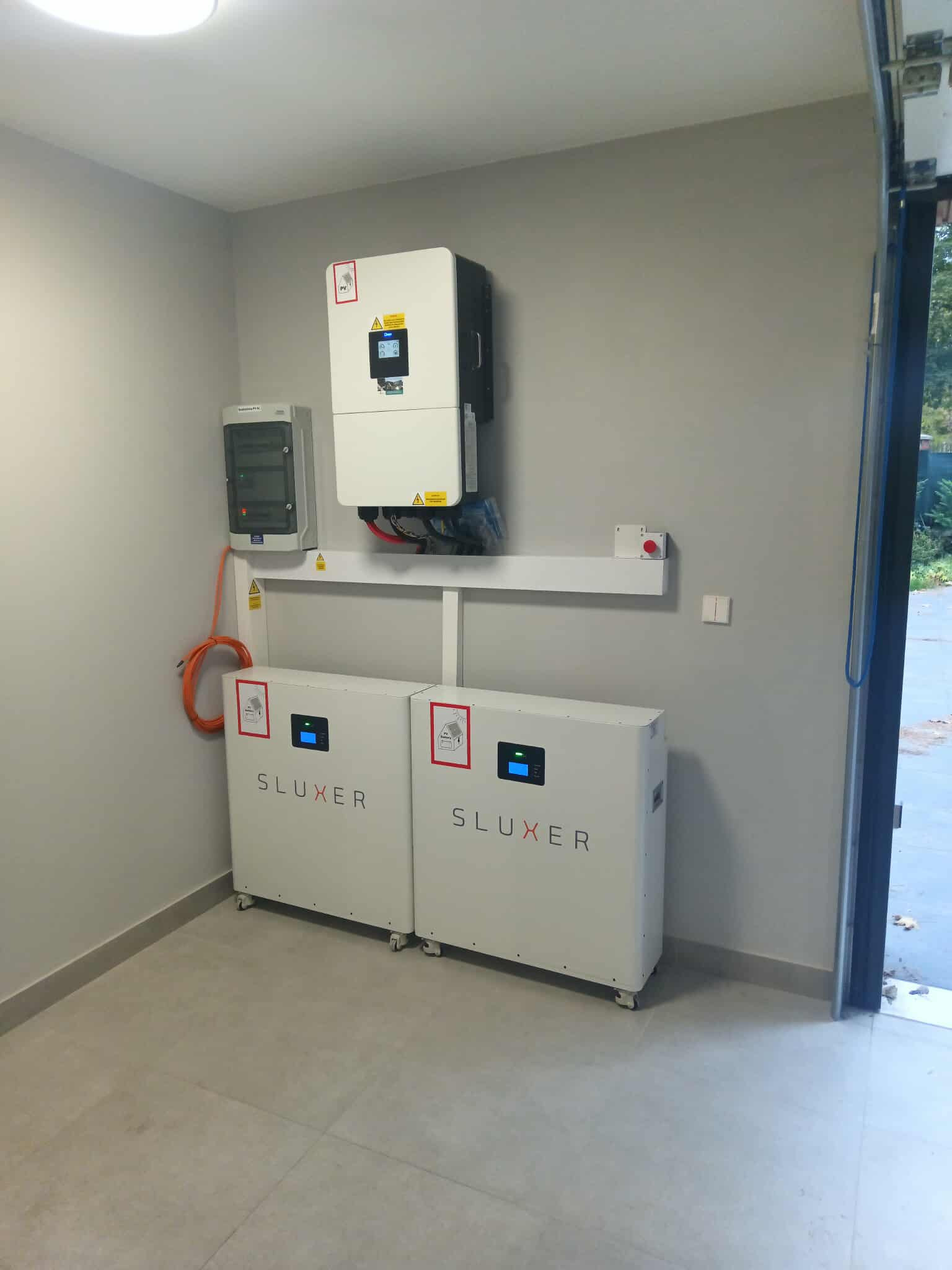 Kompletna instalacja magazynu energii Sluxer w szarym pomieszczeniu. Widoczne dwa moduły na kółkach, inwerter i rozdzielnica. Pomarańczowy kabel łączy elementy.