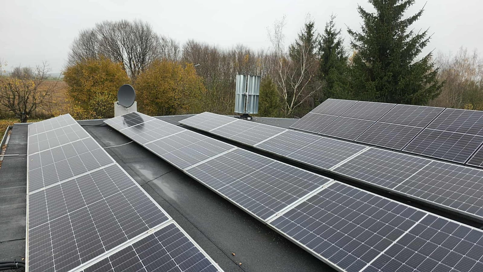 Instalacja fotowoltaiczna na dachu z turbiną wiatrową w tle. Widok z góry na panele słoneczne w jesiennej scenerii. Antena satelitarna na dachu.