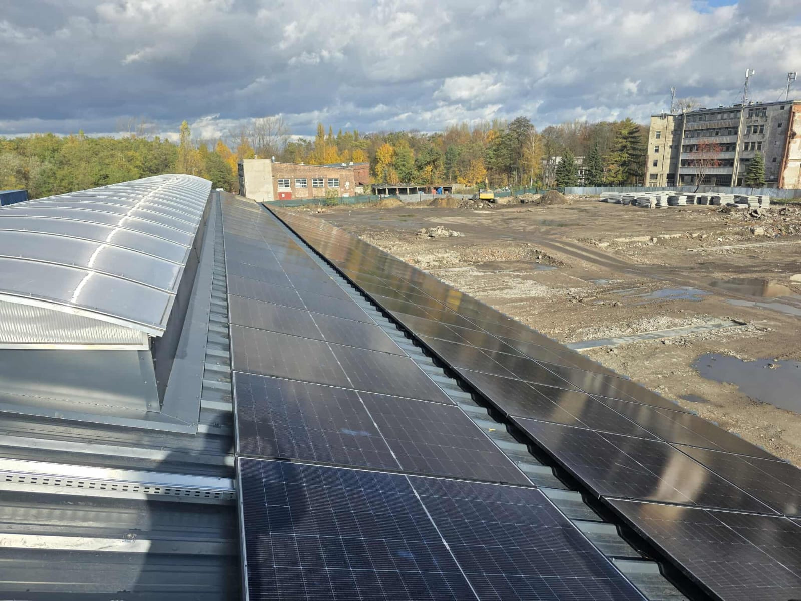 Rząd paneli słonecznych na dachu budynku z blachy trapezowej, w tle plac budowy i budynki w jesiennej scenerii pod pochmurnym niebem.