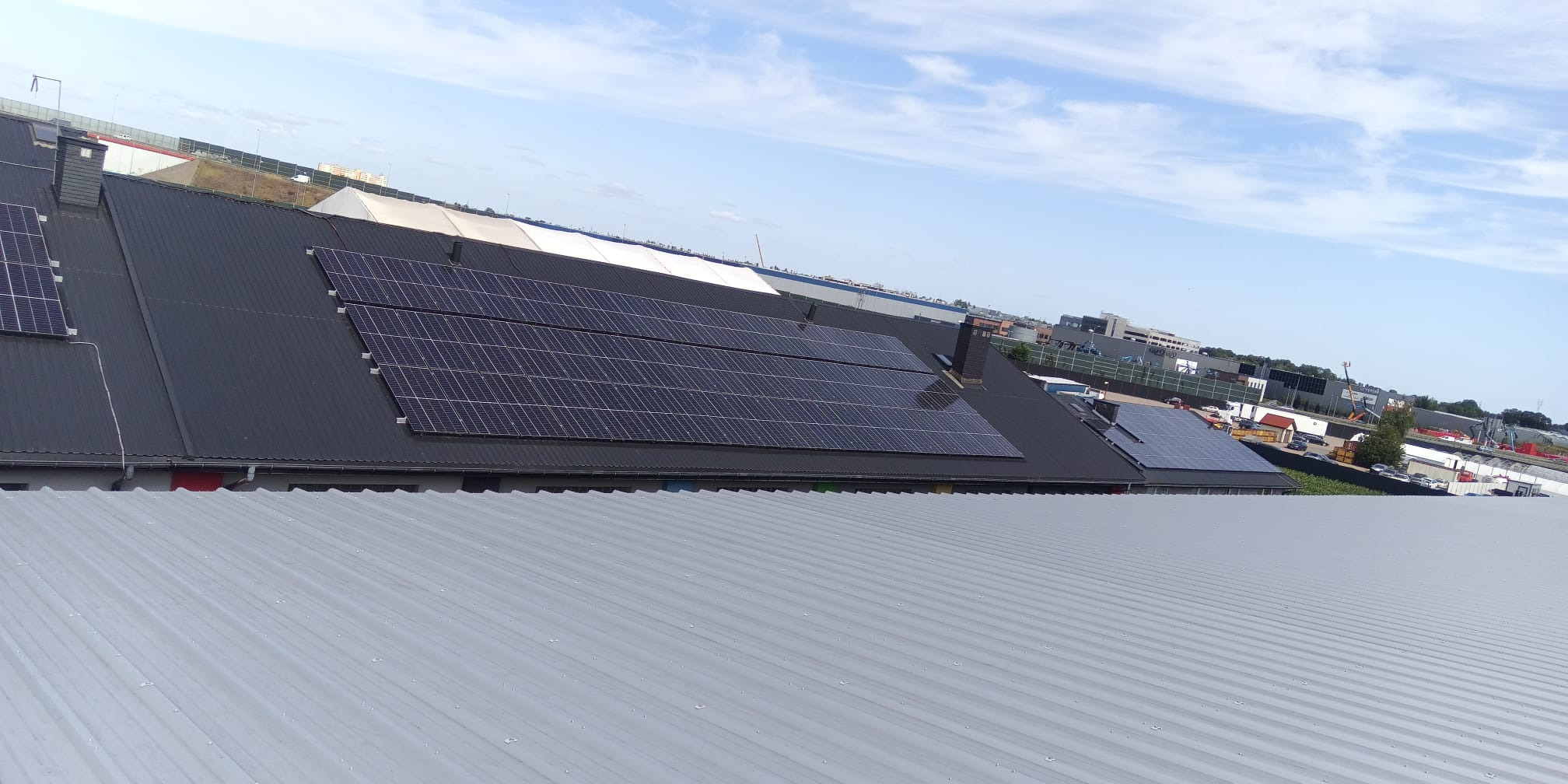 Instalacja fotowoltaiczna na dachu z blachy trapezowej, widok z góry, błękitne niebo w tle. Panele ułożone w rzędach, widoczne elementy montażowe.