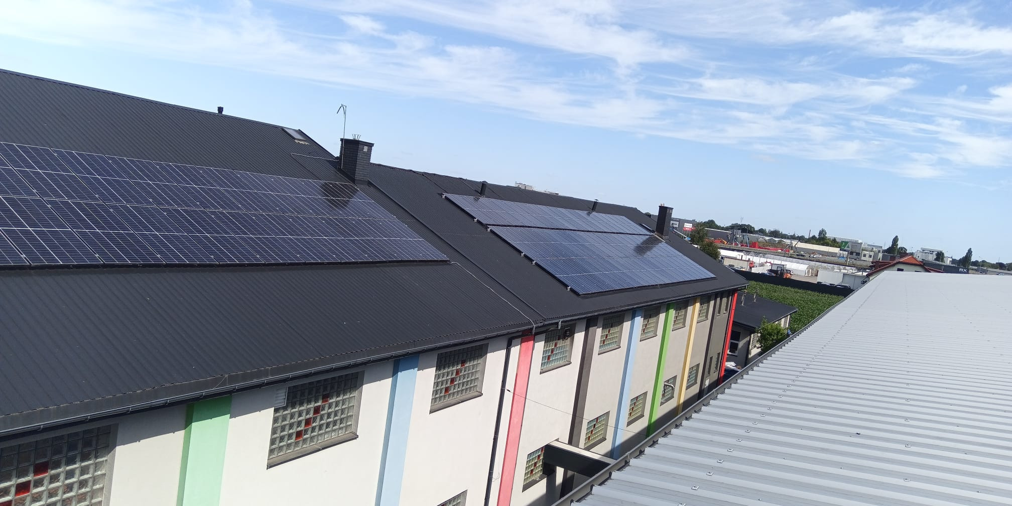 Instalacja fotowoltaiczna na dachu budynku z kolorowymi pasami. Panele słoneczne zamontowane na czarnym, metalowym dachu. Widok z góry, błękitne niebo w tle.