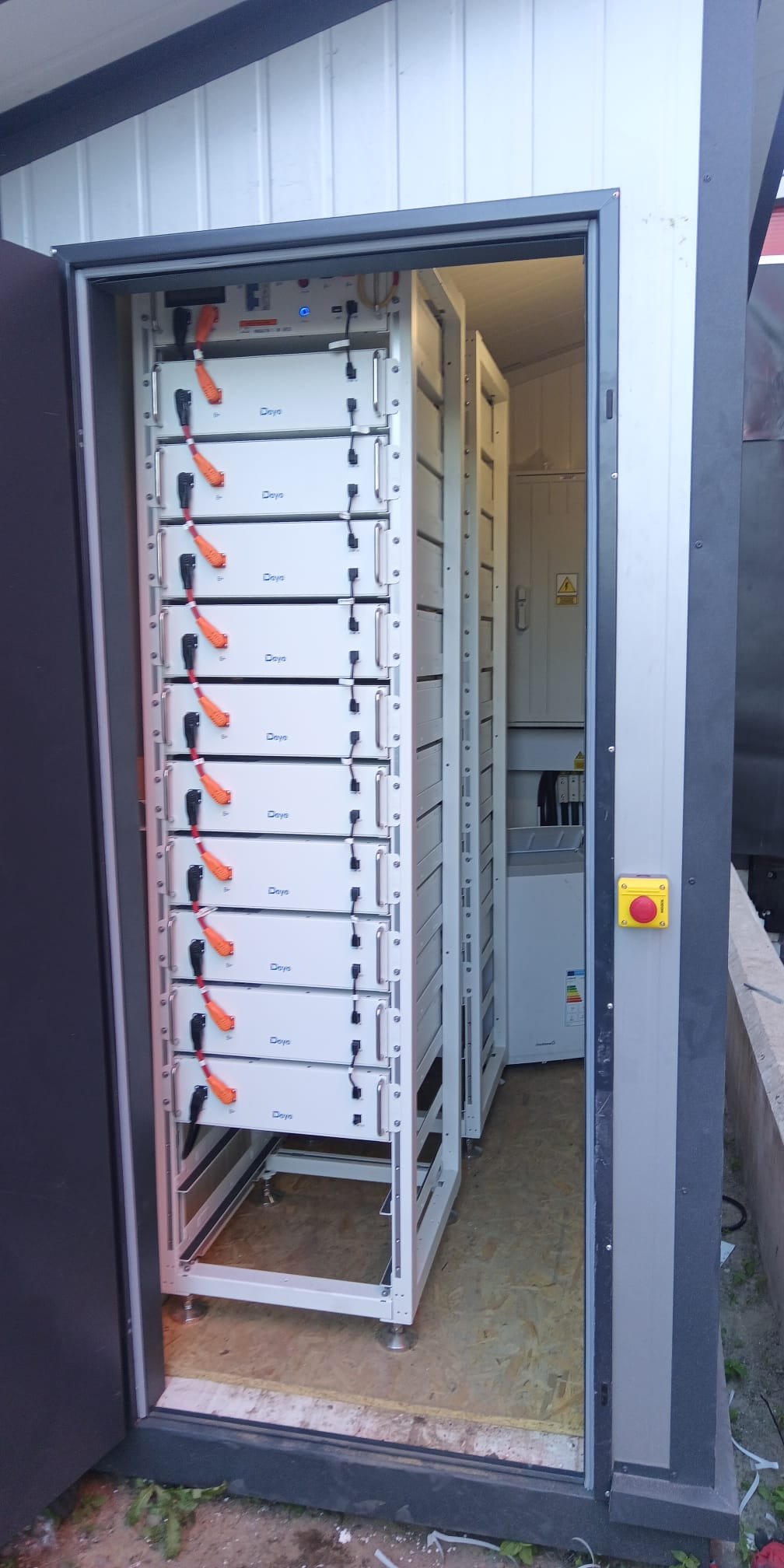 Rząd białych modułów bateryjnych Deye w metalowej szafie, widoczny przez otwarte drzwi. W tle kolejna szafa i instalacja elektryczna. Całość wewnątrz budynku.