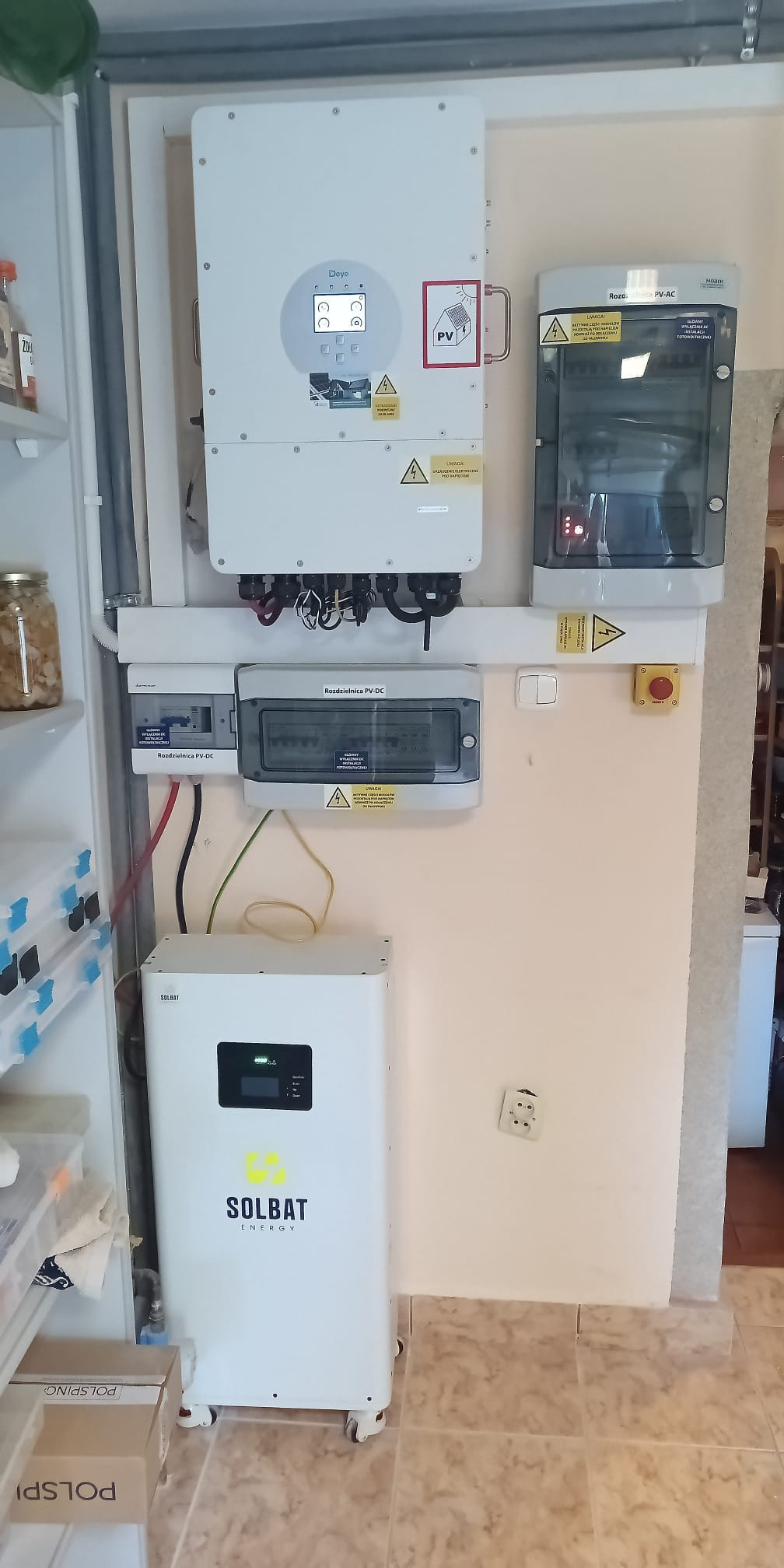 Kompletna instalacja fotowoltaiczna wewnątrz budynku: inwerter Deye, rozdzielnice PV-DC i PV-AC, oraz magazyn energii Solbat na kółkach. Widoczne okablowanie i zabezpieczenia.