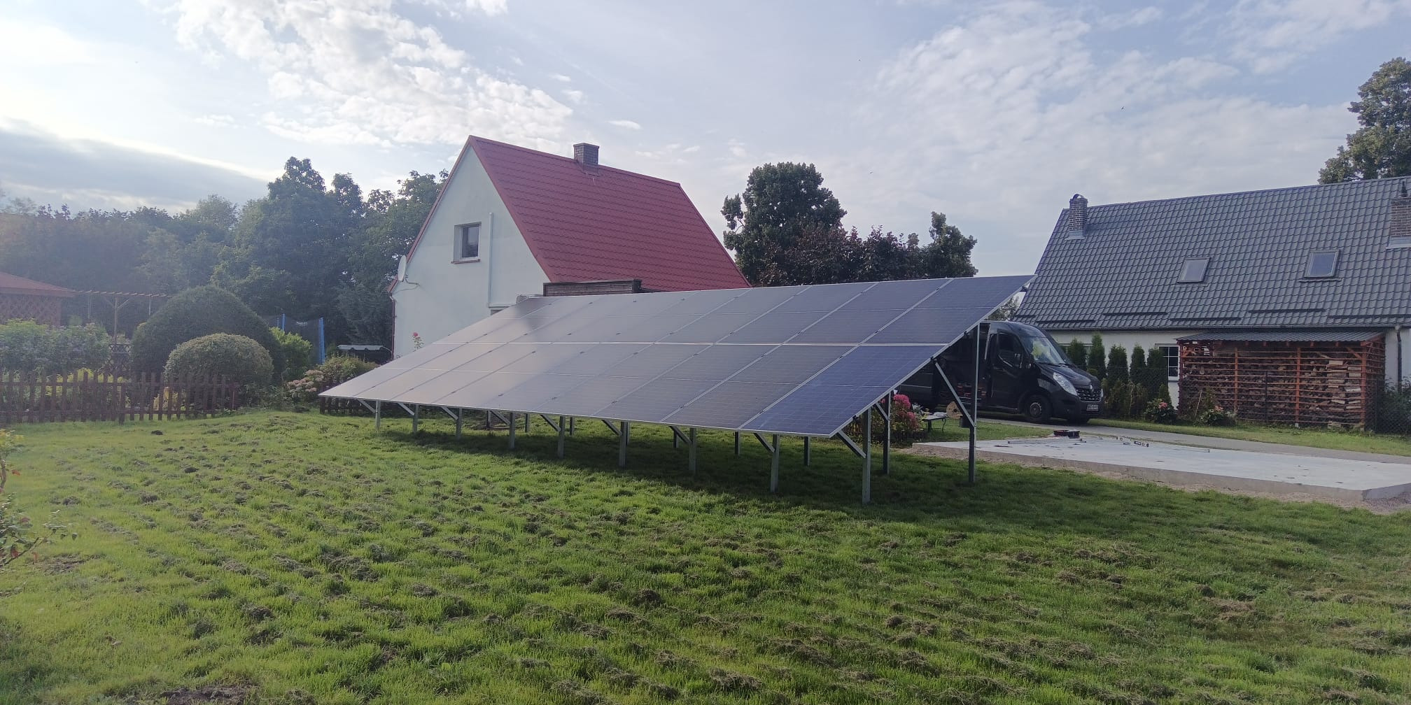 Instalacja fotowoltaiczna na trawniku przed domem z czerwonym dachem. Panele na metalowej konstrukcji, w tle zaparkowany czarny van. Słoneczny dzień.