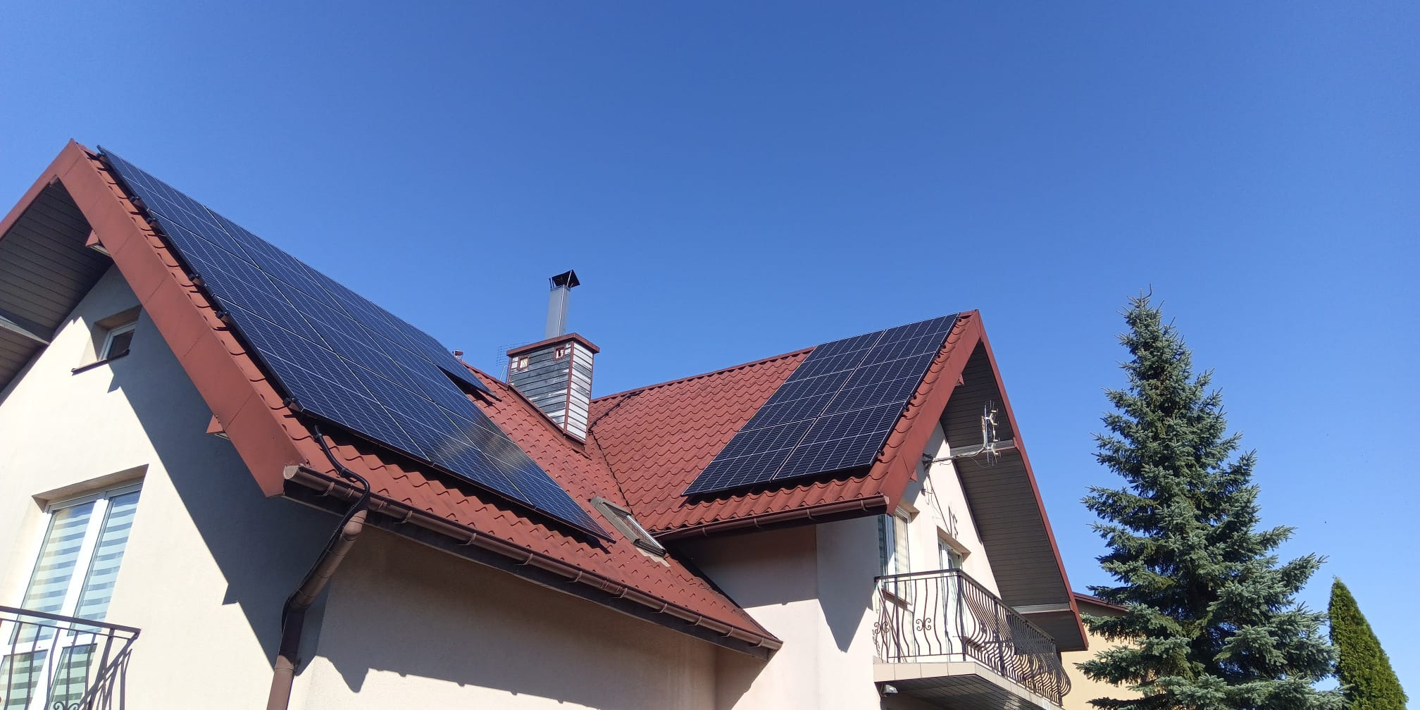 Dom jednorodzinny z panelami fotowoltaicznymi na dachu z czerwonej dachówki, komin i iglaste drzewo na tle błękitnego nieba.