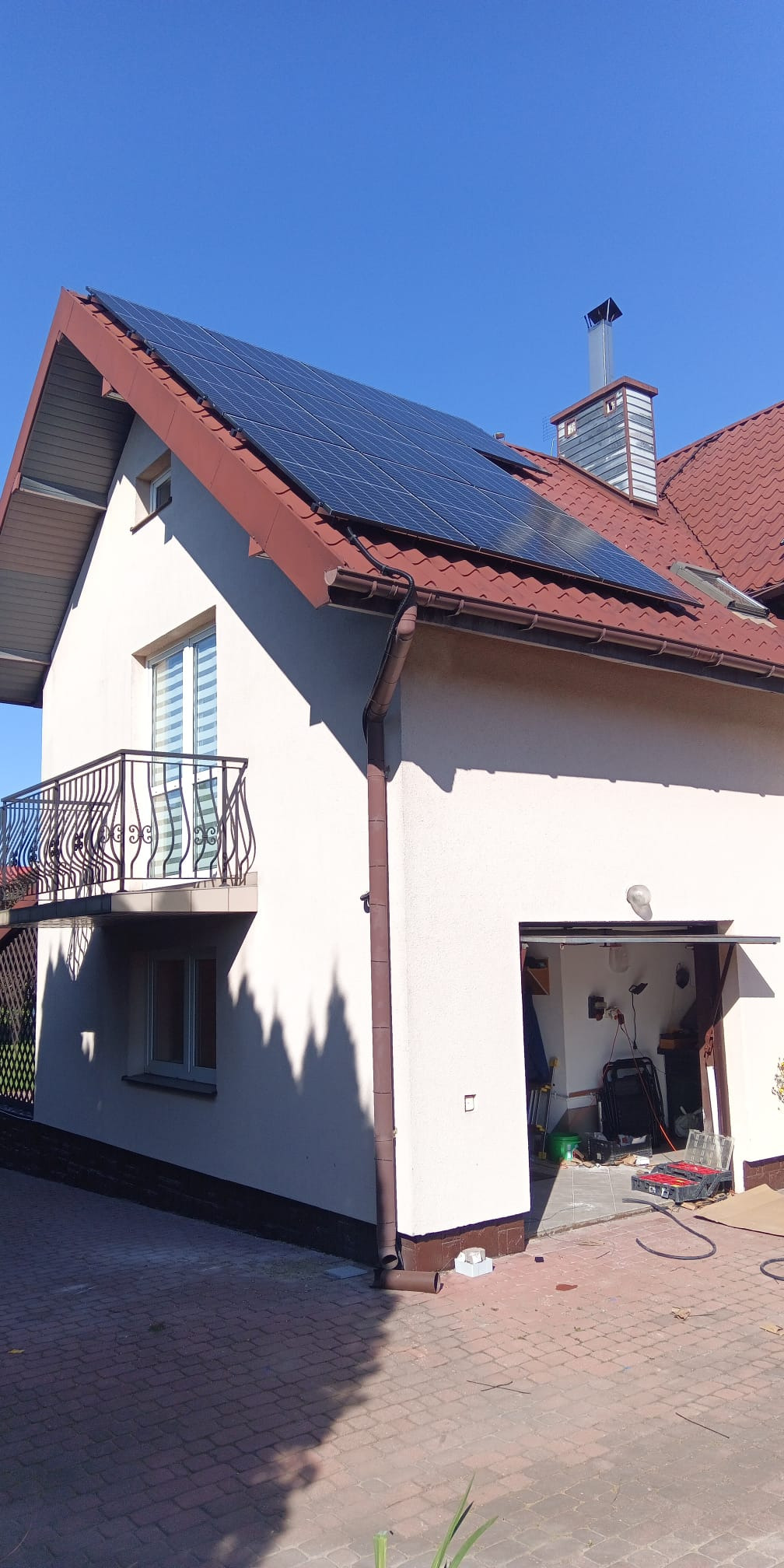Biały dom z brązowym dachem i panelami słonecznymi na dachu. Widoczny balkon z ozdobną balustradą i otwarty garaż z narzędziami w środku. Słoneczny dzień.