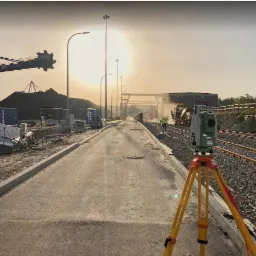 Pomiar geodezyjny nowej drogi w trakcie budowy, teodolit na statywie na pierwszym planie, w tle maszyny budowlane i robotnik w kamizelce odblaskowej.