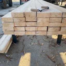 Stos świeżo przetartej tarcicy ułożony na metalowym stojaku, z leżącym na wierzchu narzędziem do obróbki drewna, w tle widoczny fragment maszyny stolarskiej i koło traktora.