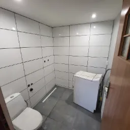 Wykończona łazienka z białymi płytkami na ścianach, szarą podłogą, toaletą i pralką, widok przez otwarte drewniane drzwi.