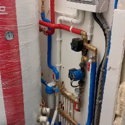 Węzeł cieplny z zasobnikiem, pompą obiegową i rozdzielaczem, rury w izolacji czerwonej i niebieskiej, regulator temperatury na ścianie, widoczne przyłącza i zawory.