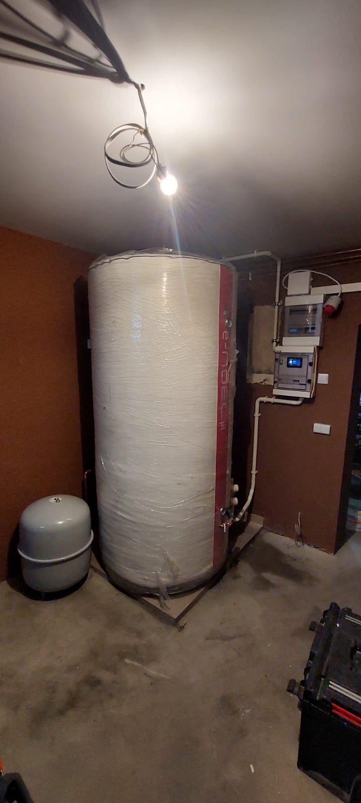 Duży, biały zbiornik na wodę owinięty folią ochronną, obok szare naczynie przeponowe, widoczne instalacje hydrauliczne i elektryczne, surowy beton na podłodze, niedokończone ściany.