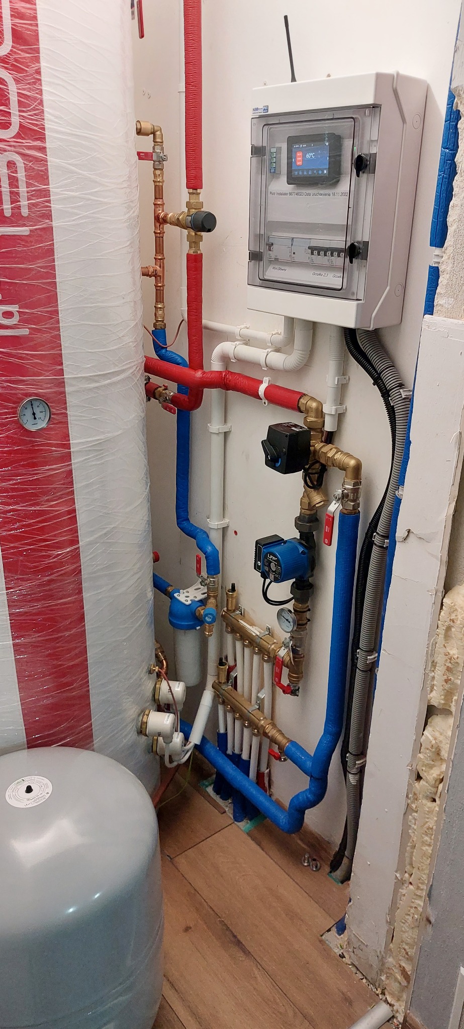 Węzeł cieplny z zasobnikiem, pompą obiegową i rozdzielaczem, rury w izolacji czerwonej i niebieskiej, regulator temperatury na ścianie, widoczne przyłącza i zawory.