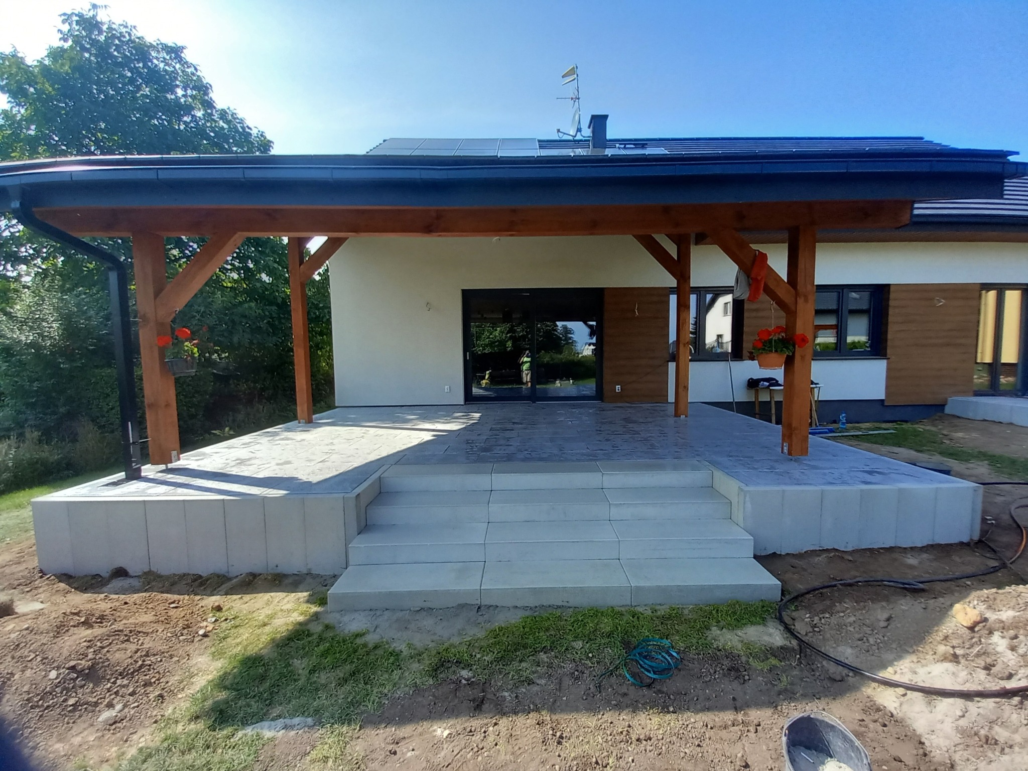 Nowoczesny taras z szarej kostki brukowej, schody z kostki, drewniana pergola z kwiatami, dom z panelami solarnymi w tle.