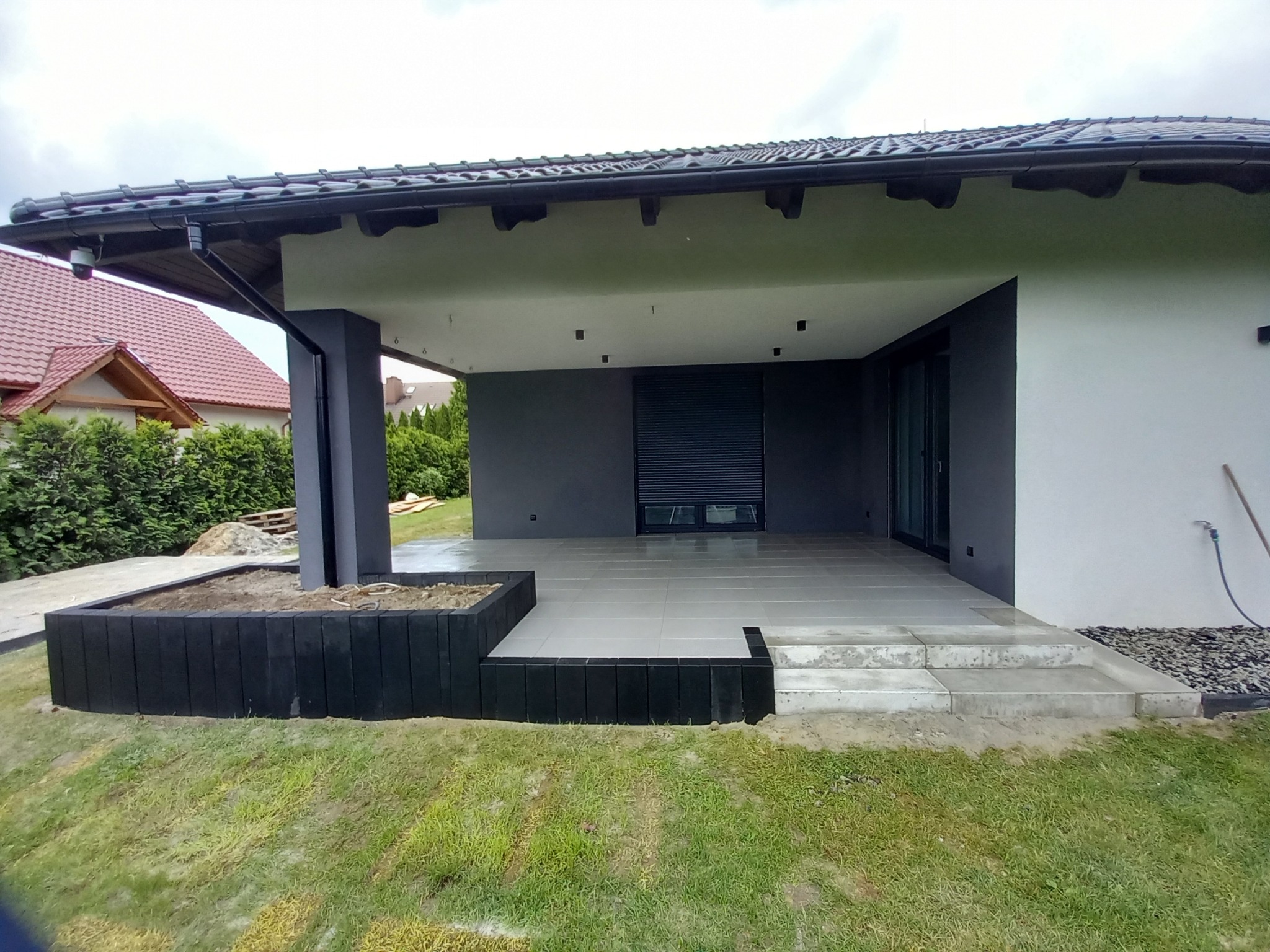 Nowoczesny taras z jasnoszarą kostką, czarnymi obrzeżami i schodami, widok na dom z ciemnymi elementami wykończenia.