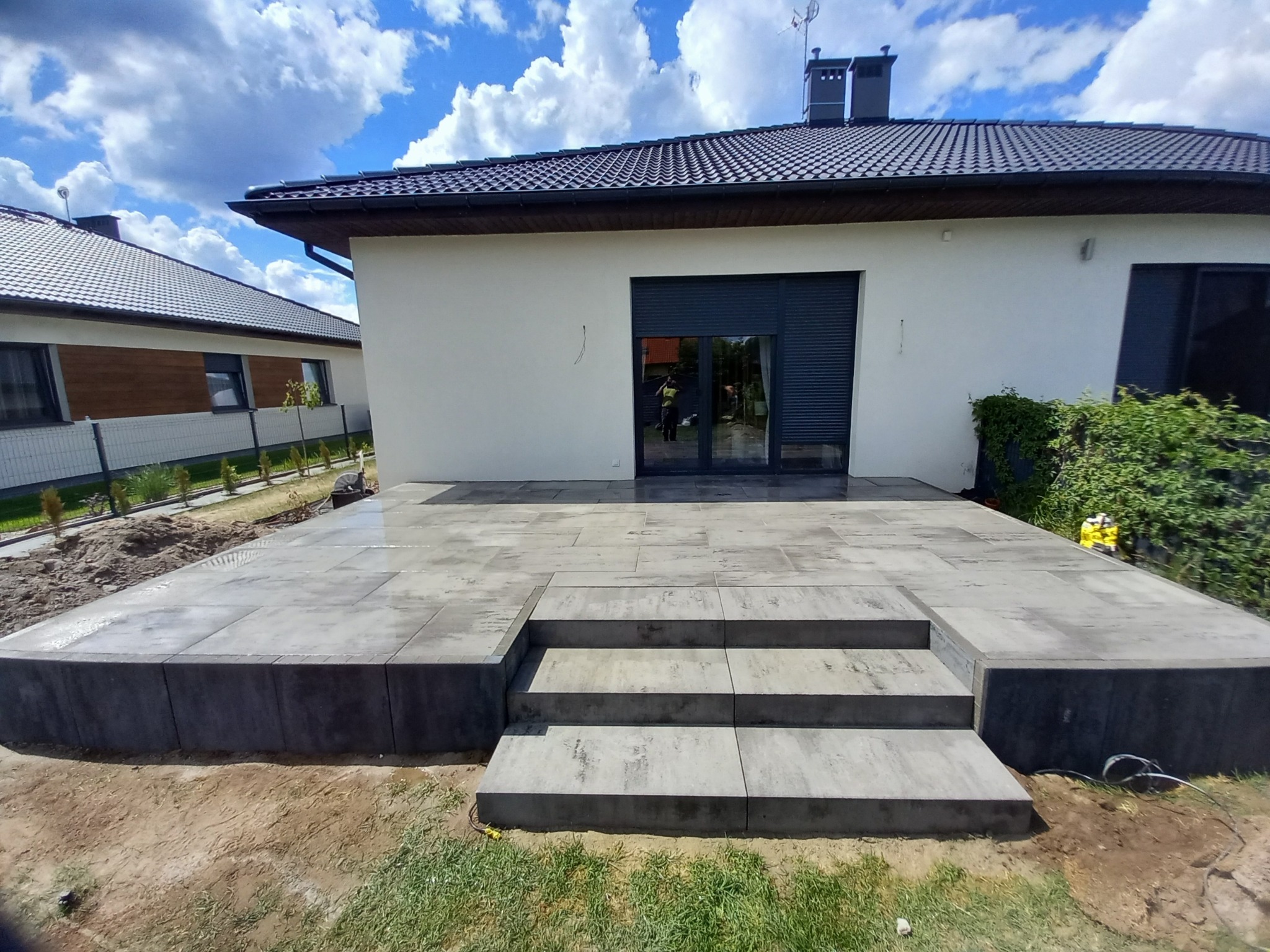 Nowoczesny taras z szarych płyt betonowych z trzema schodami, przylegający do białego domu z czarną stolarką okienną i drzwiami, na tle błękitnego nieba z chmurami.