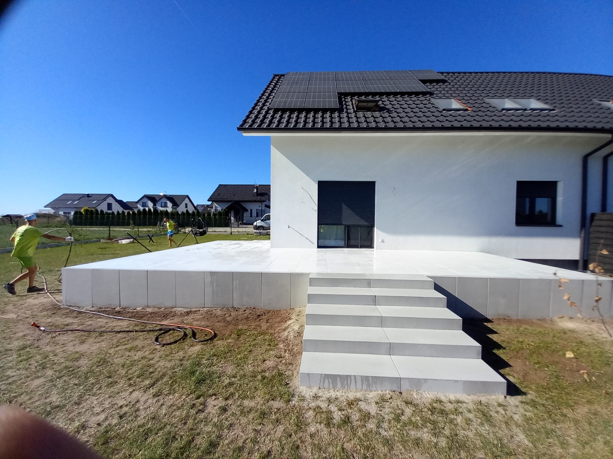 Nowoczesny taras z jasnoszarej kostki brukowej, z widocznymi schodami i fragmentem domu z panelami słonecznymi na dachu, w trakcie mycia pod ciśnieniem przez pracownika w zielonej koszulce.