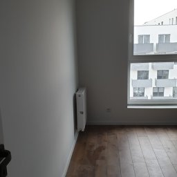 Wolter-BUD - Pusty pokój po remoncie z drewnianą podłogą, świeżo pomalowanymi szarymi ścianami, białym grzejnikiem i oknem z widokiem na nowoczesny budynek.
