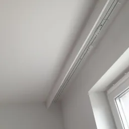 Białe wnętrze z zamontowaną szyną karniszową pod sufitem, widoczne mocowania i okno z widokiem na zewnątrz.