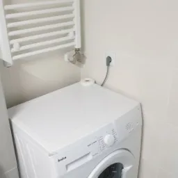 Biała pralka Amica w narożniku łazienki z grzejnikiem drabinkowym na ścianie wyłożonej jasnymi płytkami.