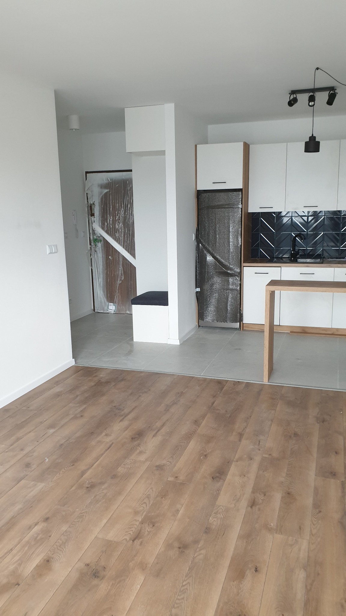 Wnętrze mieszkania w trakcie remontu: widoczna podłoga z paneli, fragment kuchni z czarną mozaiką na ścianie, zabezpieczone folią drzwi i szafka.