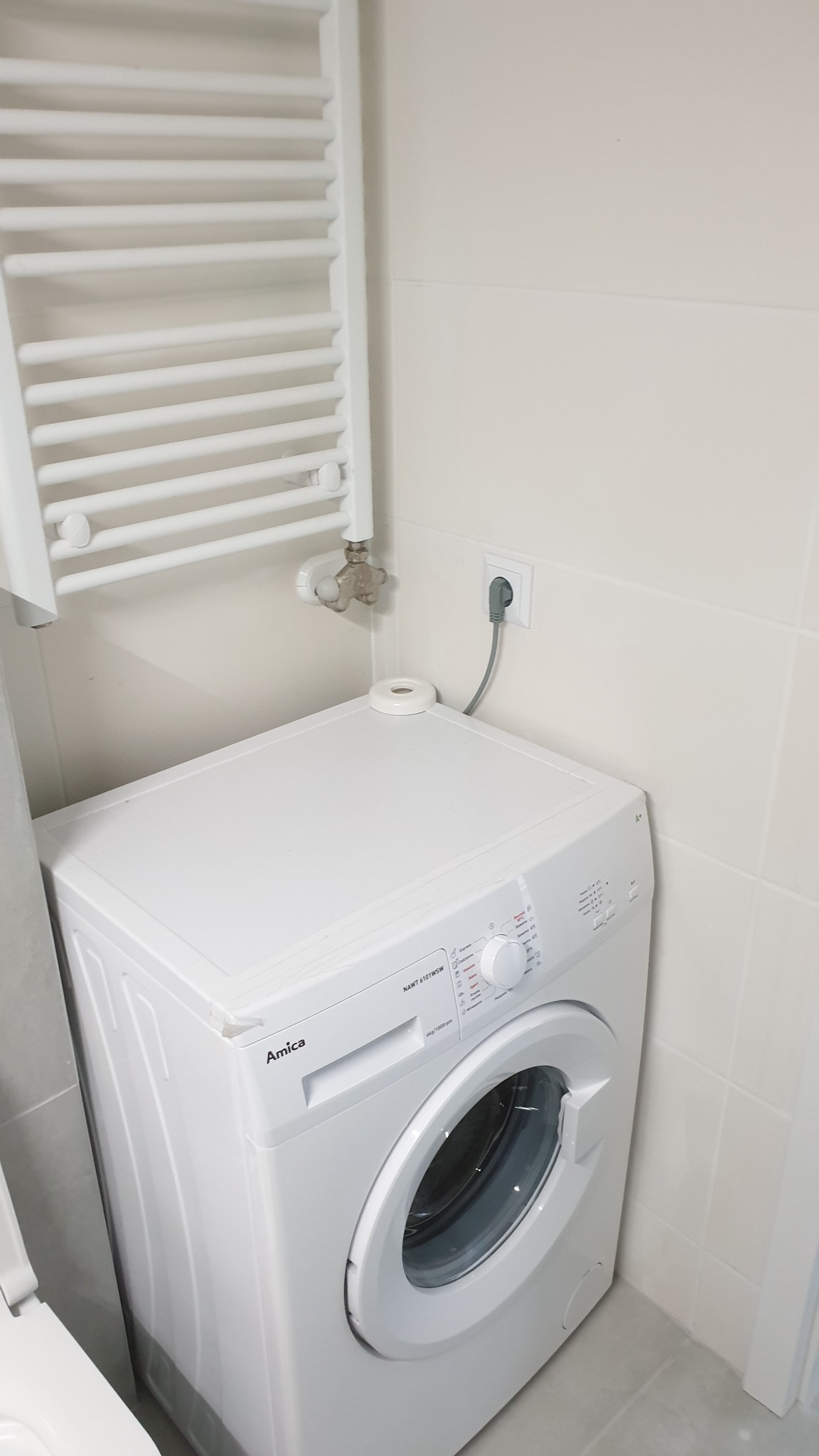 Biała pralka Amica w narożniku łazienki z grzejnikiem drabinkowym na ścianie wyłożonej jasnymi płytkami.
