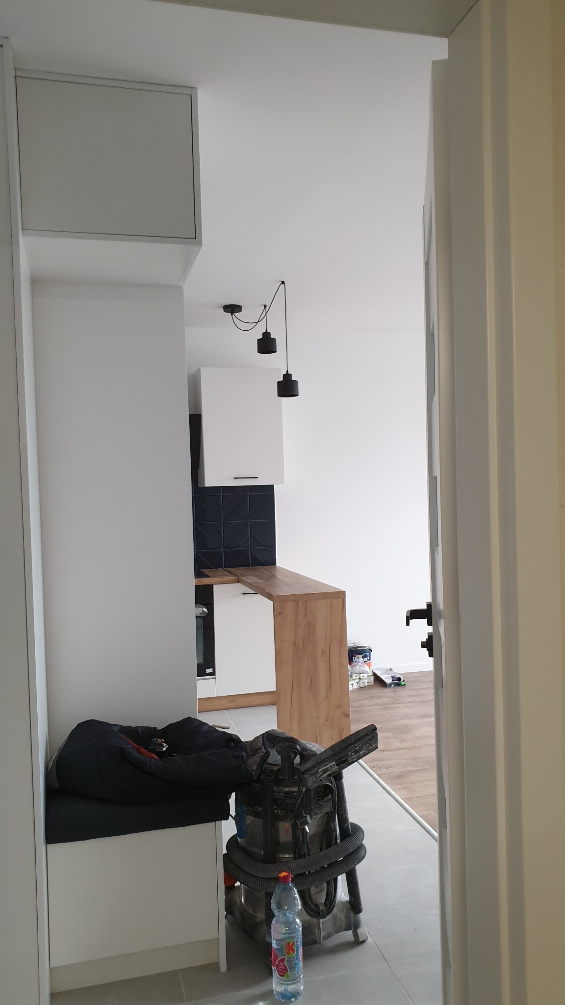 Wnętrze mieszkania w trakcie wykańczania, widoczny odkurzacz przemysłowy, fragment zabudowy kuchennej z drewnianym blatem oraz czarne płytki na ścianie.