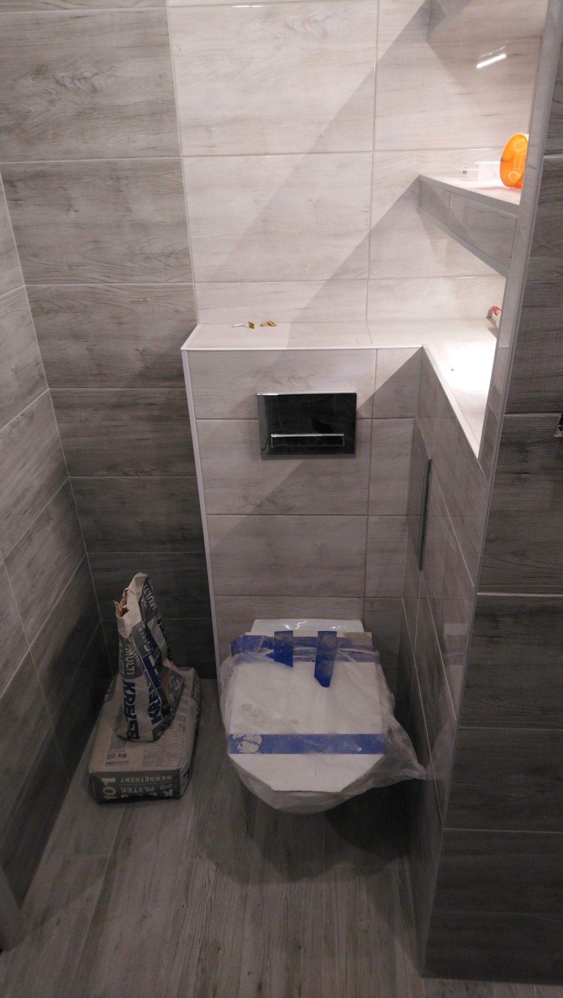 Montaż podtynkowego WC z szarą mozaiką imitującą drewno na ścianach, widoczny worek z klejem do płytek i zabezpieczona folią miska sedesowa.