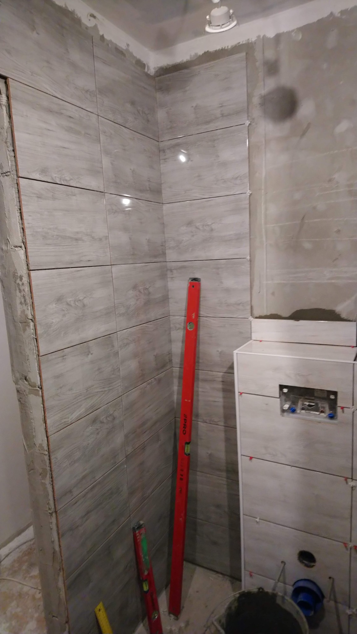 Ściana łazienki w trakcie układania płytek imitujących drewno, widoczne poziomice pionowe, zabudowa stelaża WC z częściowo ułożonymi płytkami, szary grunt na ścianie obok.