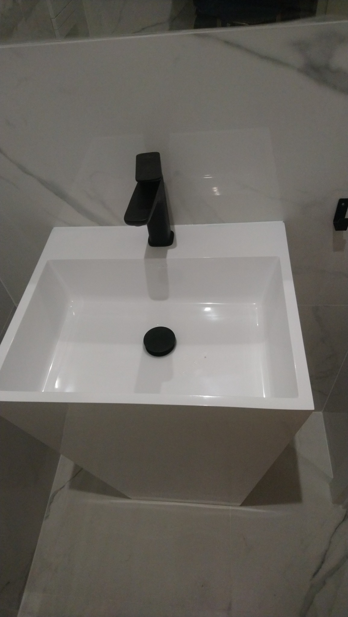 Nowoczesna, biała umywalka nablatowa z czarną, minimalistyczną baterią i odpływem, zamontowana na tle płytek imitujących marmur.