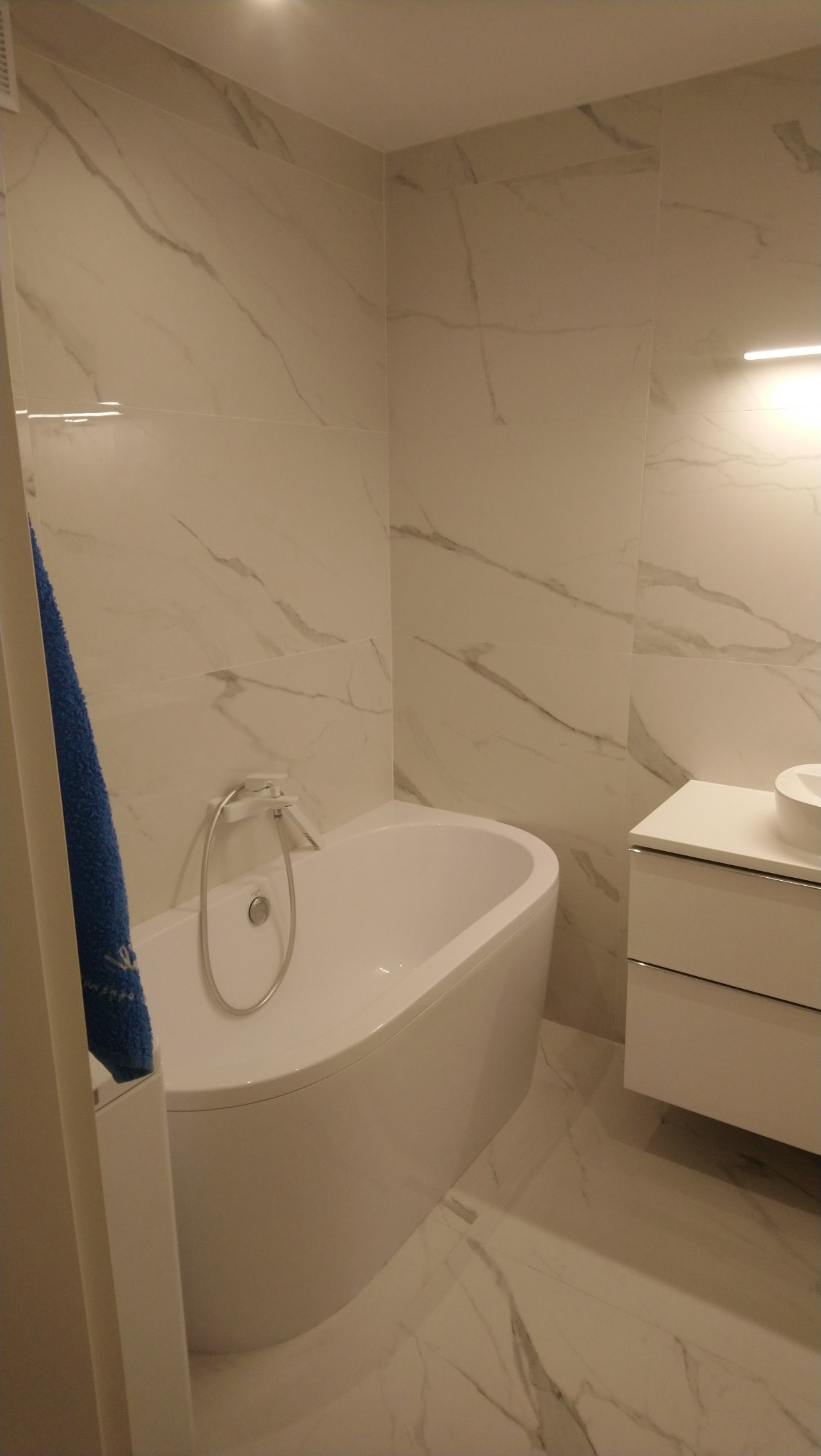 Nowoczesna łazienka z białą wanną, umywalką z szafką i marmurowymi płytkami na ścianach i podłodze, z niebieskim ręcznikiem na wannie.
