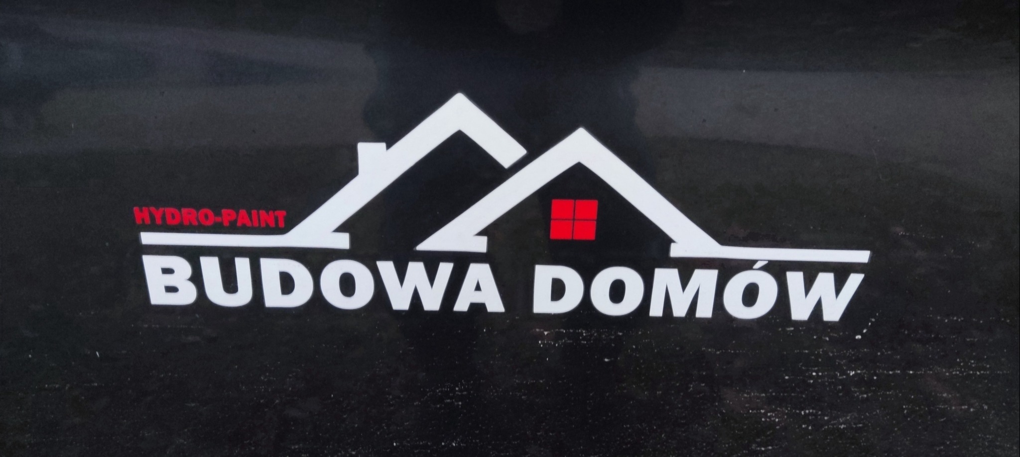Logo firmy Hydro-Paint z białym symbolem domu z czerwoną szybą i napisem 'Budowa Domów' na czarnym tle.