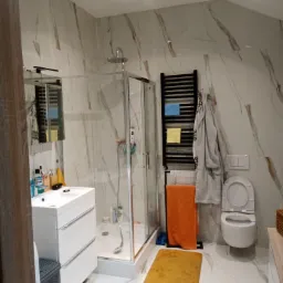 Nowoczesna łazienka z kabiną prysznicową, umywalką z szafką, grzejnikiem drabinkowym z ręcznikami i marmurowymi płytkami na ścianach i podłodze.