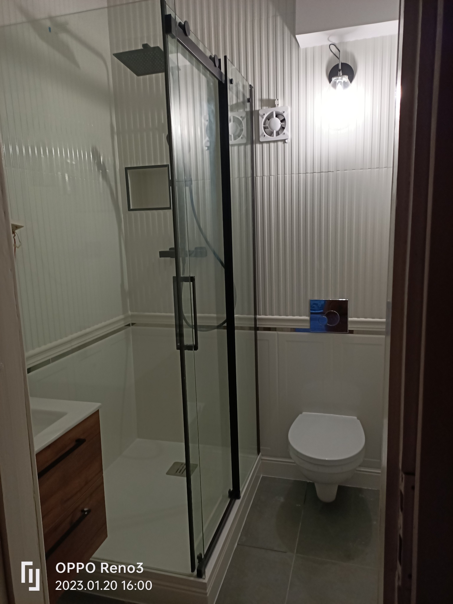 Nowoczesna łazienka z kabiną prysznicową typu walk-in z czarnymi ramami, toaletą i umywalką z drewnianą szafką, wykończona białą boazerią pionową i szarą podłogą z płytek.