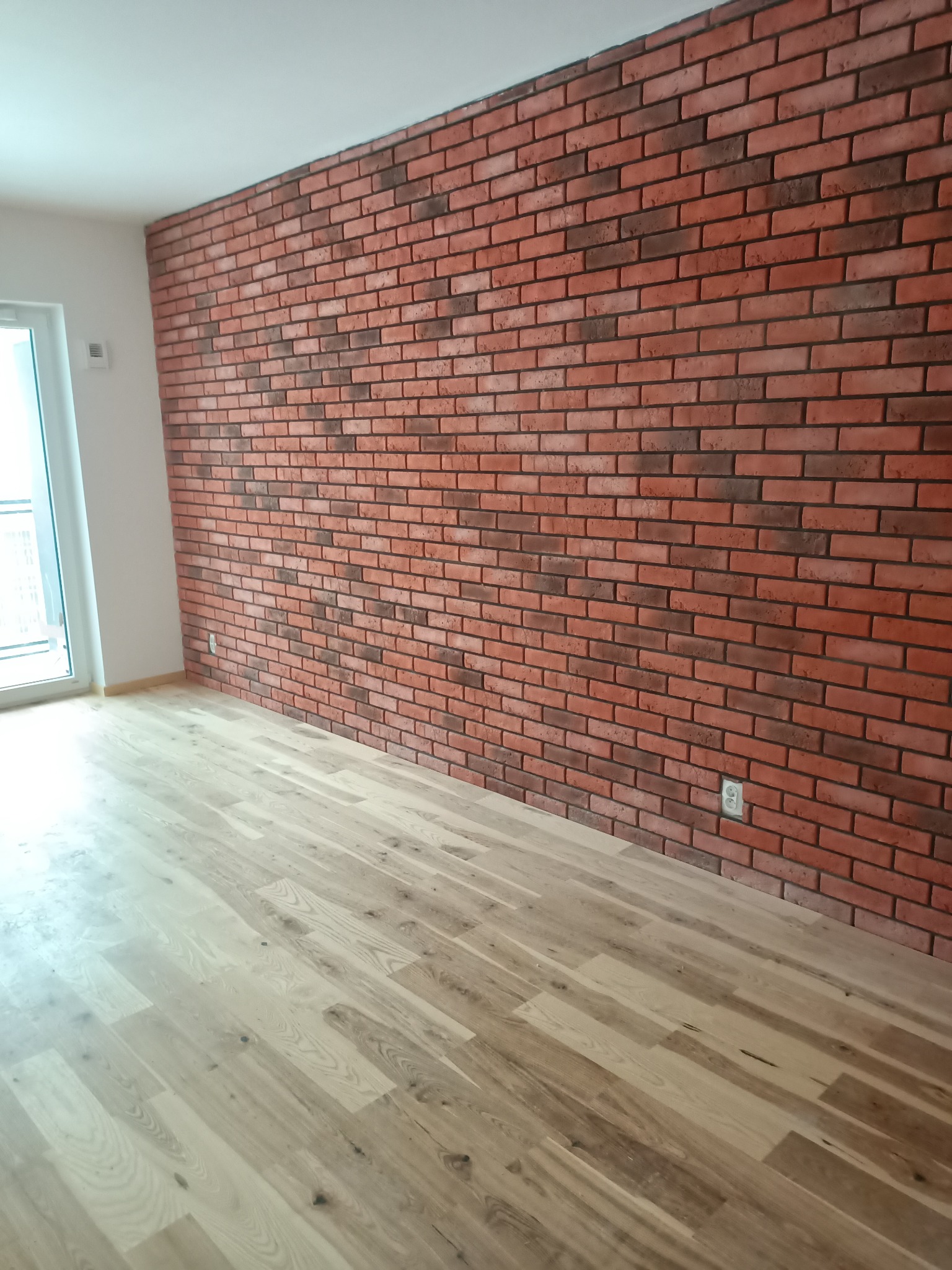 Ściana wykończona czerwoną cegłą dekoracyjną, jasna drewniana podłoga z paneli, białe ściany i okno w nowym mieszkaniu.