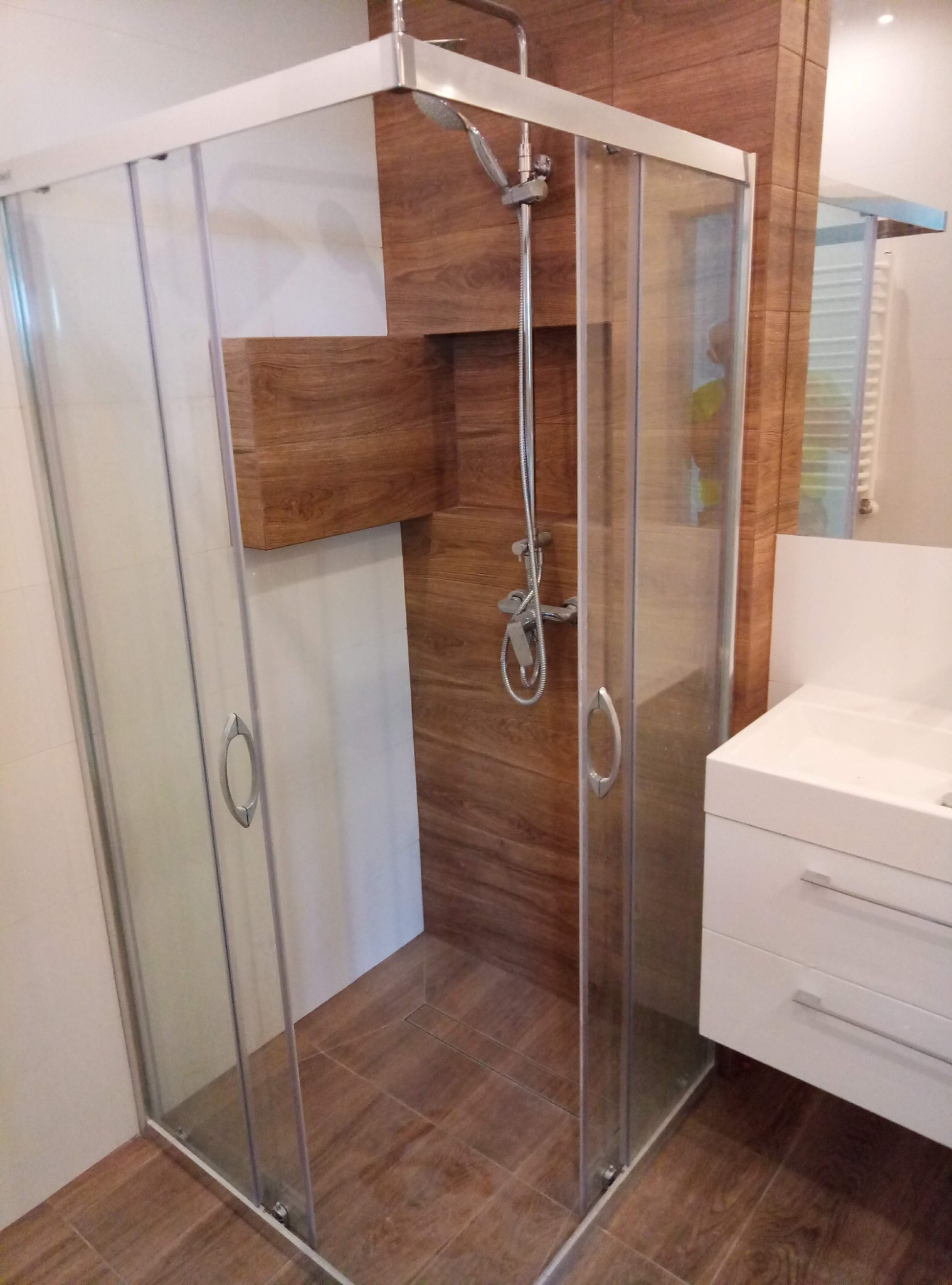Nowoczesna kabina prysznicowa z przesuwnymi drzwiami i chromowanymi uchwytami, wykończona płytkami imitującymi drewno, z widoczną białą szafką umywalkową i grzejnikiem drabinkowym w tle.
