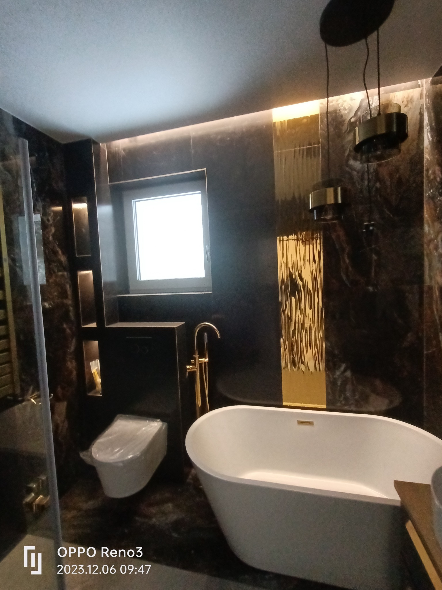 Nowoczesna łazienka z wanną wolnostojącą, toaletą podwieszaną i ciemnymi marmurkowymi płytkami, akcentowana złotymi elementami oświetlenia i armatury.