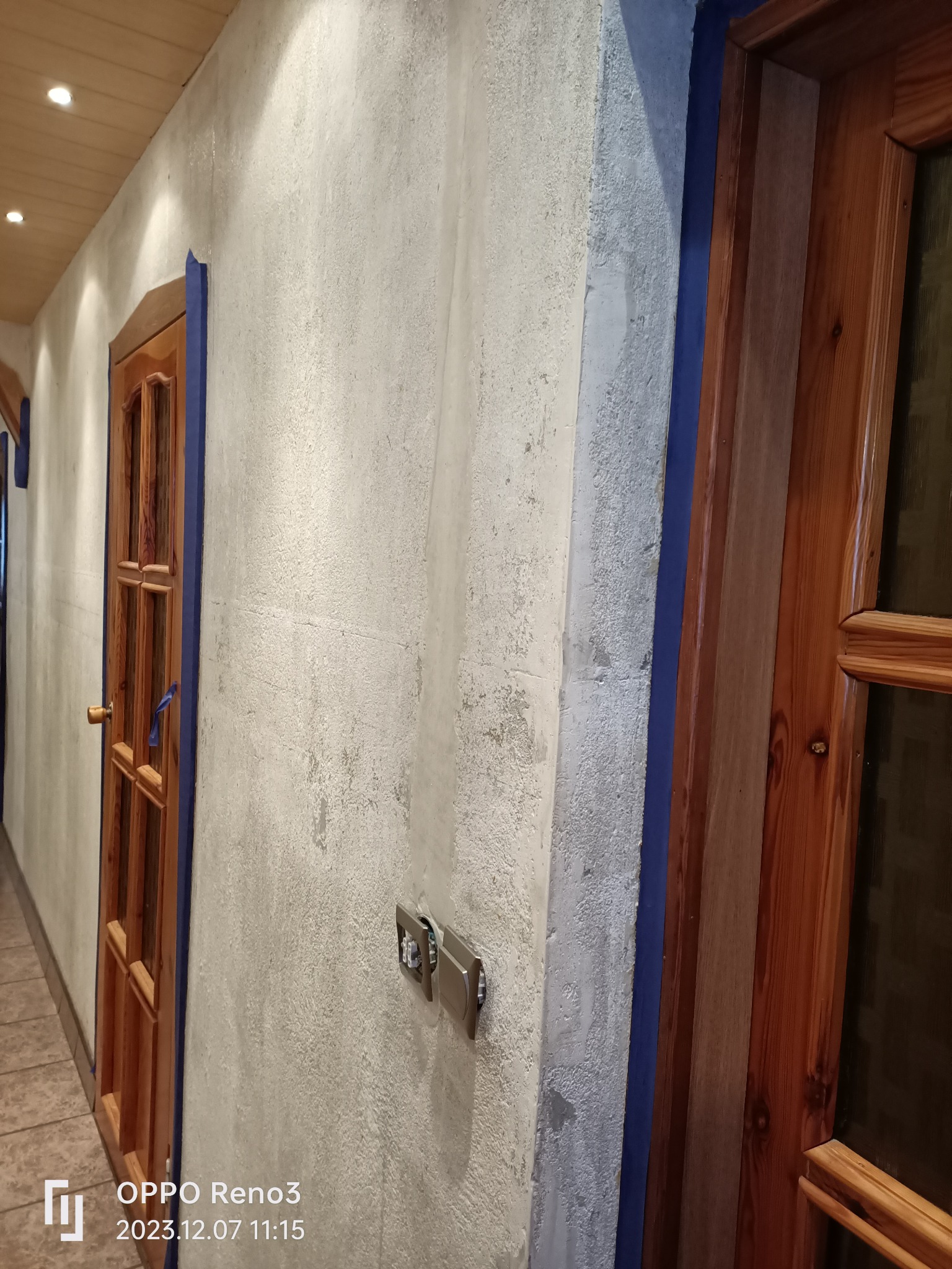 Ściana z nałożoną szarą gładzią, zabezpieczona niebieską taśmą malarską wokół drewnianych framug drzwi i kontaktów elektrycznych, widoczne nierówności faktury.