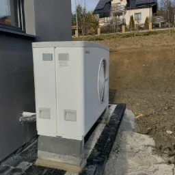 Biała pompa ciepła marki Stiebel Eltron zamontowana na betonowym fundamencie przy szarej ścianie budynku, w tle dom z panelami słonecznymi na dachu.