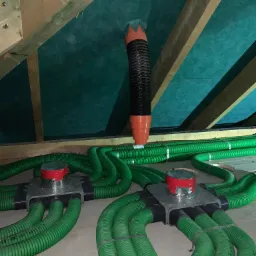 Zielone, elastyczne przewody wentylacyjne połączone z metalową skrzynką rozdzielczą, widoczne poddasze z izolacją i fragmentem rury wentylacyjnej w kolorze czarno-pomarańczowym.