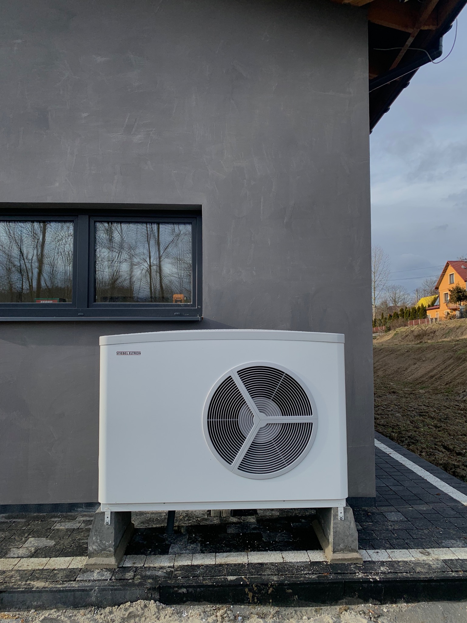 Biała jednostka zewnętrzna z wentylatorem, zamontowana na betonowych podstawach przy szarej ścianie domu z oknem, na tle zaoranego pola i domów.