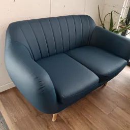 Niebieska, dwuosobowa sofa z pionowymi przeszyciami na oparciu, ustawiona na drewnianej podłodze, z roślinami w tle przy oknie.