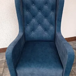 Niebieski, tapicerowany fotel typu uszak z pikowanym oparciem na tle białej ściany i kostki brukowej.