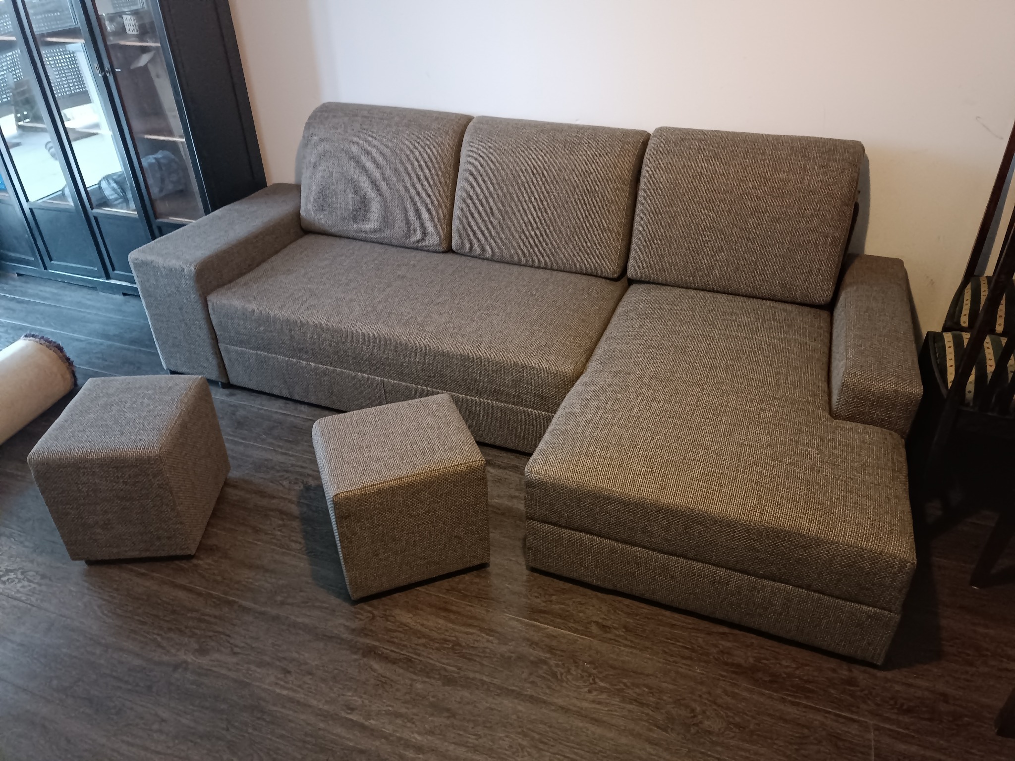 Szary narożnik z dwoma pufami na drewnianej podłodze, widoczny fragment przeszklonej szafy w tle.